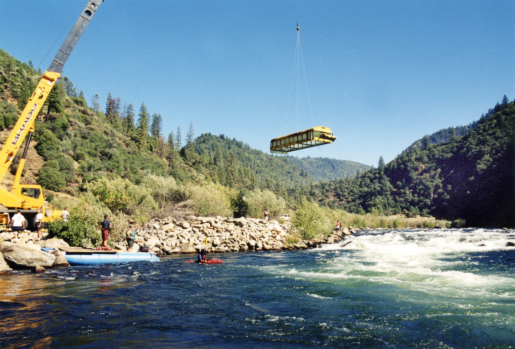 A yellow bus hanging from crane wires over the North Fork of the American River.