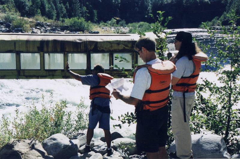 Three people in orange life vests watch as half a bus is lowered into a river.