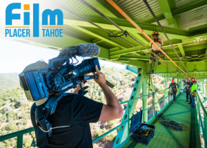 An image of a man in black holding a large, black video camera standing on a green bridge filming others. 