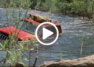 Breakdown image of a car in a river with a play button overlay.